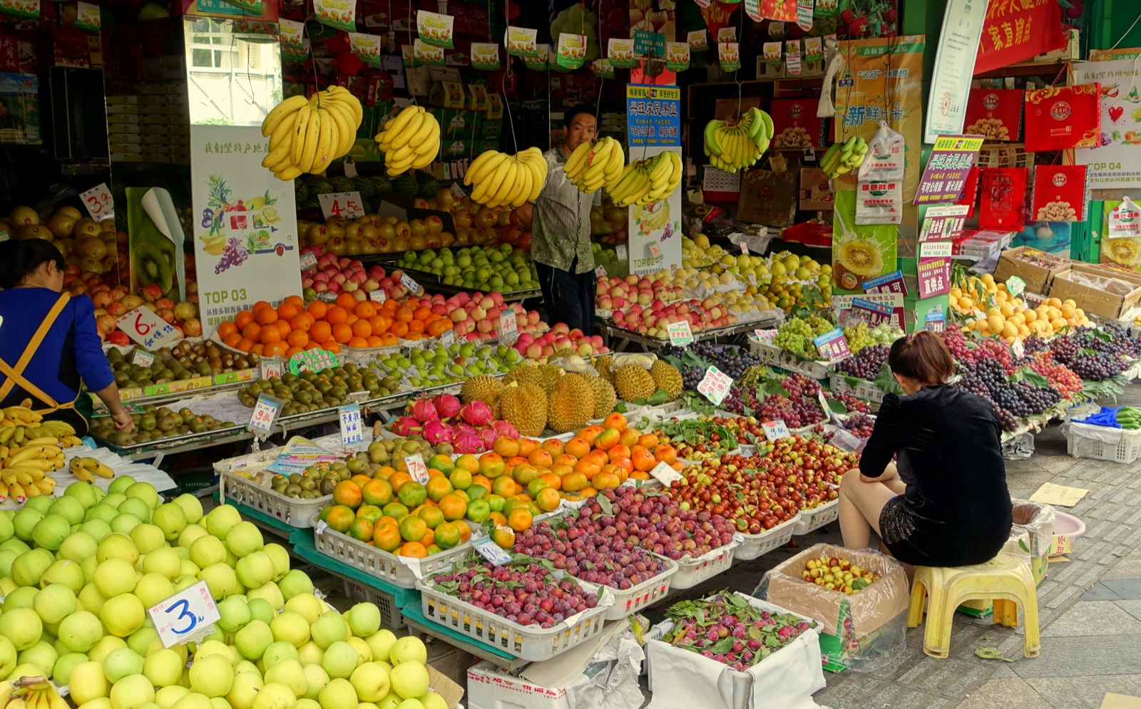 Fruta expuesta en cestas frente a una tienda de Chengdu, con racimos de plátanos colgados, carteles de precios y personas entre los puestos.