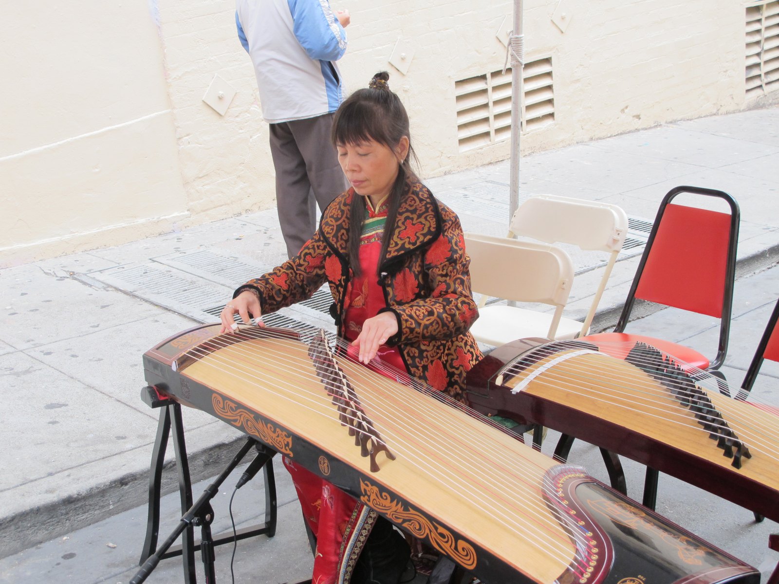 A seated musician plays a guzheng on a pavement, with a second instrument and folding chairs beside her.
