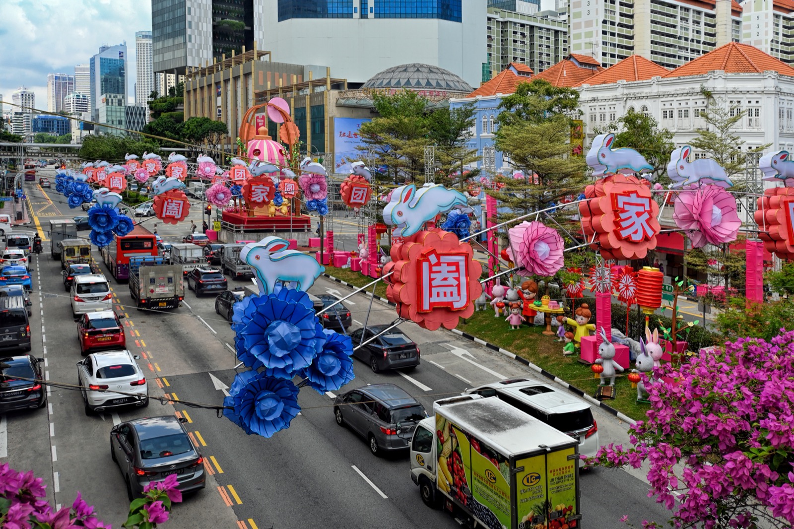 Rabbit and flower lantern decorations span a busy Singapore street, with buses, cars, shophouses and taller buildings behind them.