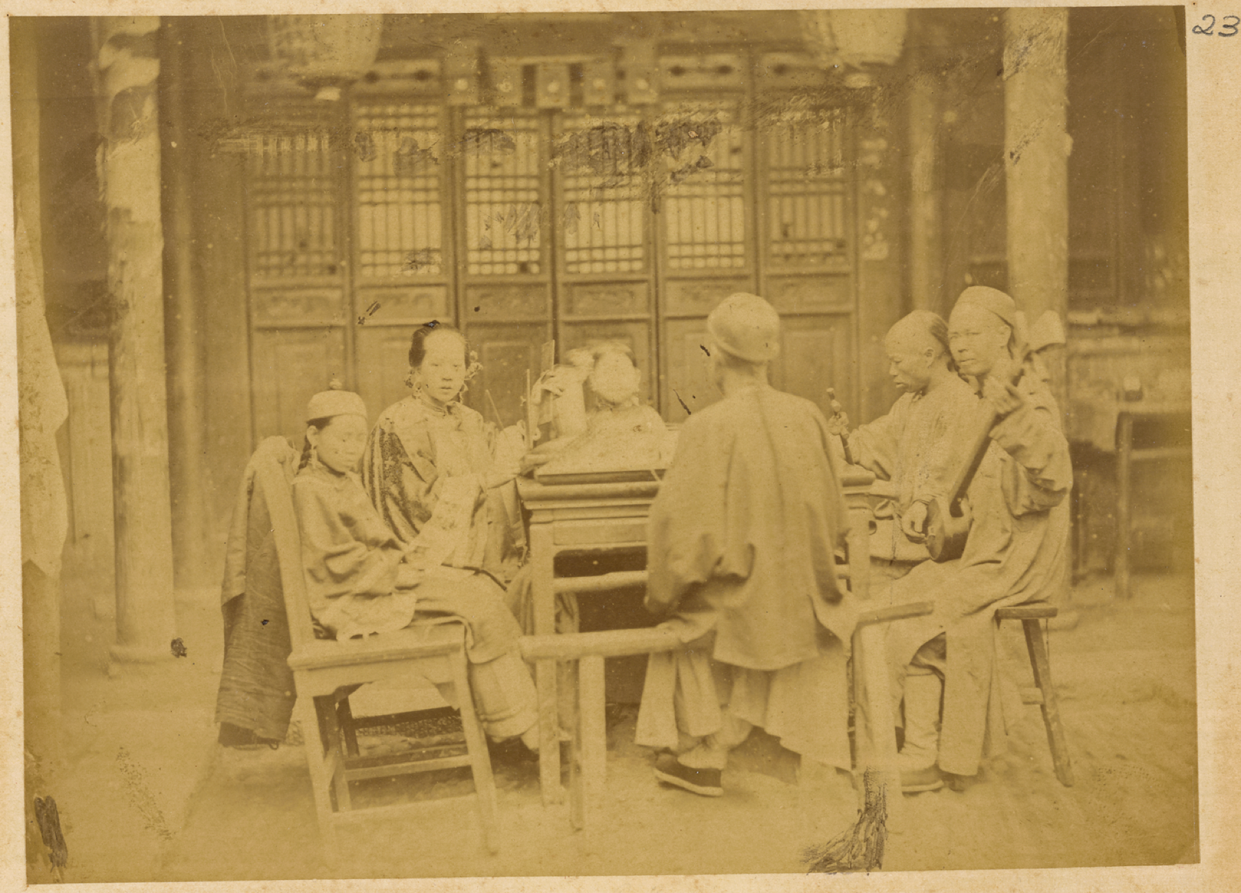 A family gathers at a table while musicians perform in China, photographed in 1874–75.