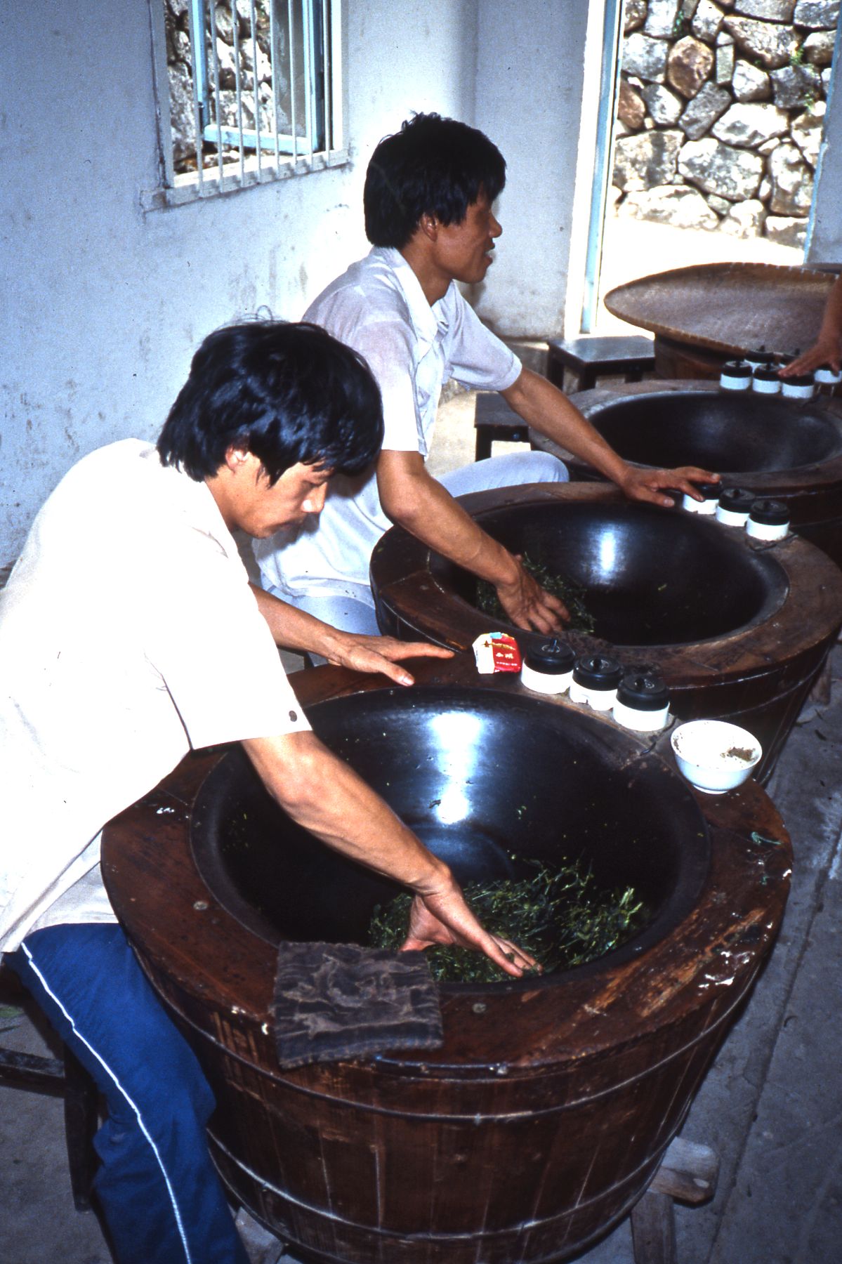 Two workers heating tea leaves by hand in large metal pans